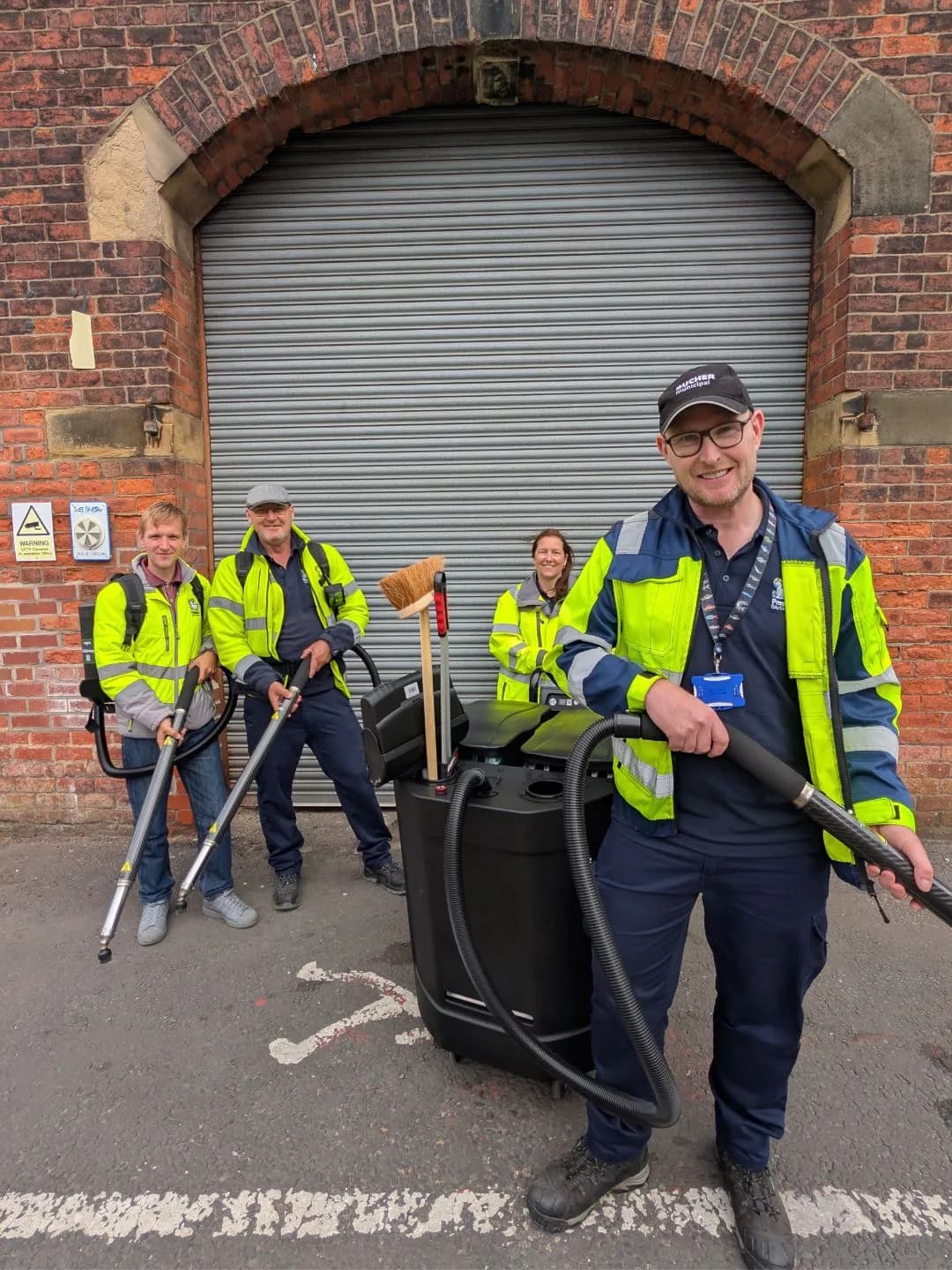 Four people in high-visibility jackets and work trousers stand with cleaning equipment, including long poles and a large black vacuum cleaner, in front of a brick building with a large metal roller door.