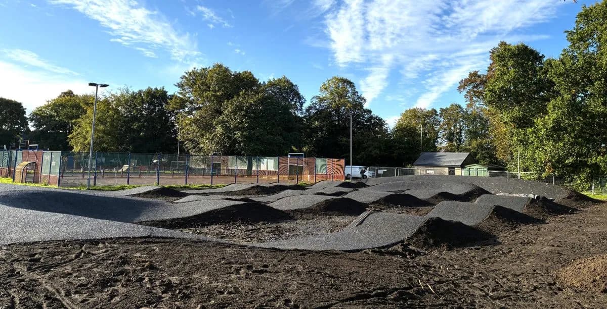 A newly constructed black tarmac pump track with various mounds and curves, surrounded by disturbed dark soil and green trees under a bright blue sky with wispy clouds.