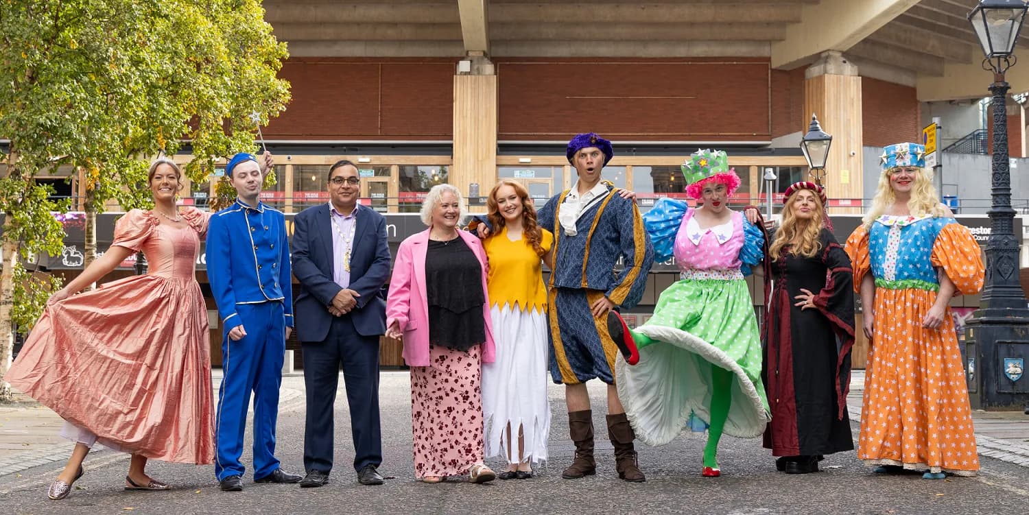 A group of ten people, some dressed in vibrant theatrical costumes and others in business attire, stand together on a paved outdoor area in front of a modern building with a tree on the left.