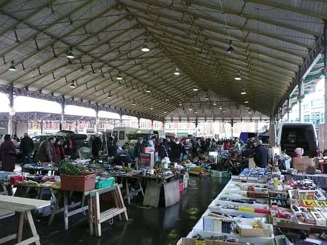 A wide shot of a bustling indoor flea market with numerous vendors, tables laden with goods, and shoppers browsing under a high, vaulted ceiling with exposed metal trusses and overhead lighting.